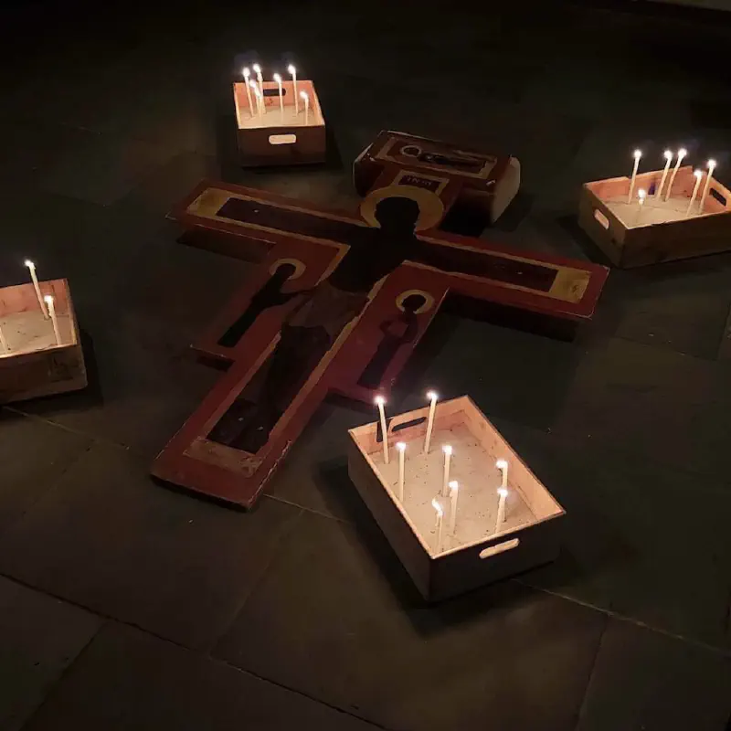 Taizé-Kreuz, das auf dem Boden der Kirche liegt, umgeben von vier mit Sand gefüllten Holzkästen, in denen jeweils einige brennende Kerzen stecken.