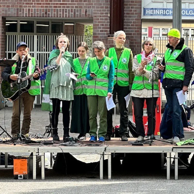 Sieben Menschen in grünen Westen stehen auf einer Bühne und singen. Eine Person hat eine Gitarre. Eine Person hält ein Mikrofon.