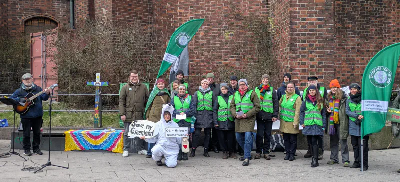 Gruppenbild der Organisator:innen der Klimaandacht. Die meisten Personen tragen grüne Warnwesten. Eine Person ist als Eisbär verkleidet. Die Gruppe steht neben einem mit Schöpfungsmotiven bemalten Kreuz. Eine Person hat eine Gitarre umgehängt.