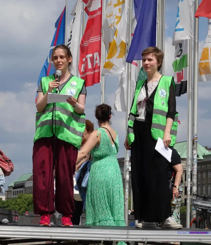 Zwei Frauen in grünen Warnwesten mit Christians4Future-Aufdruck halten eine Rede